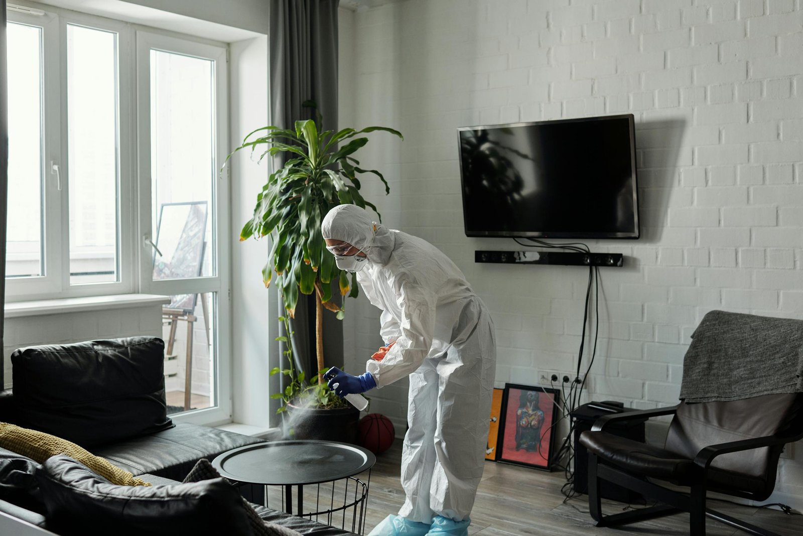 Worker in PPE disinfecting a living room, ensuring safety from viruses.