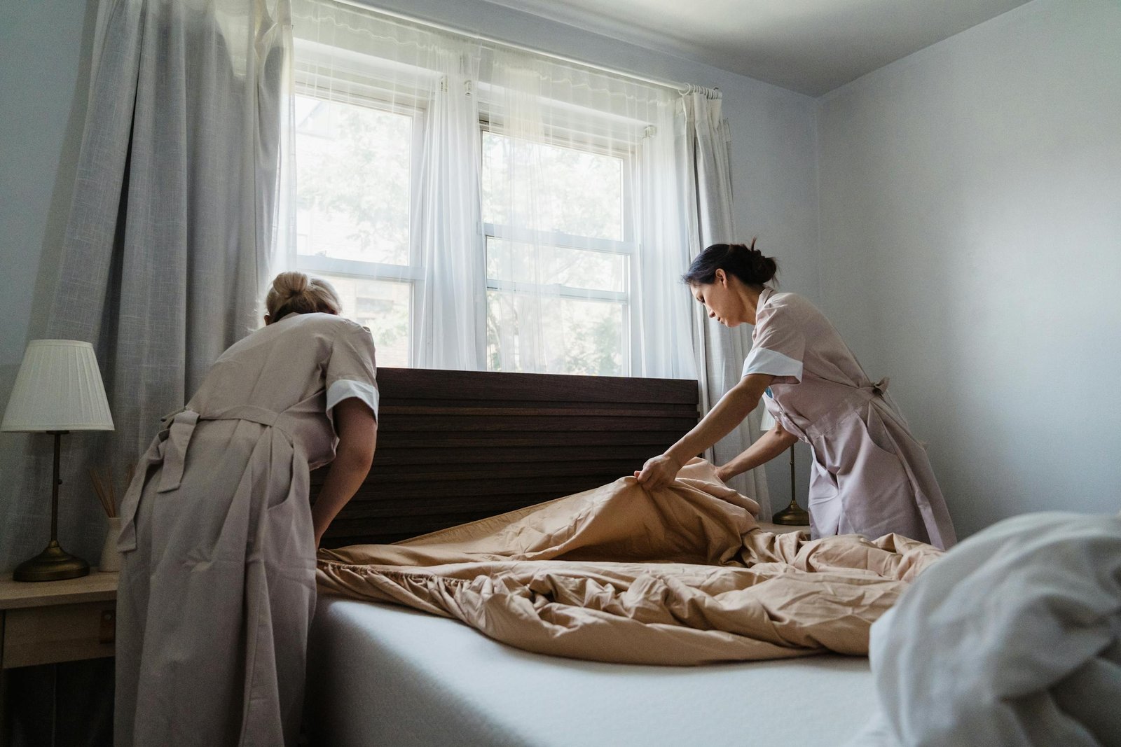 Two housekeepers making a bed in a sunlit bedroom, showcasing cleanliness and attention to detail.